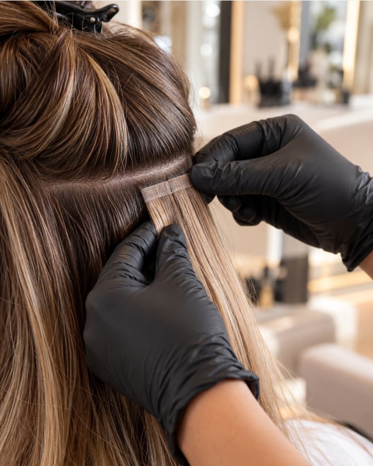 Close-up of a tape-in hair extension weft being positioned against natural brunette hair with caramel highlights